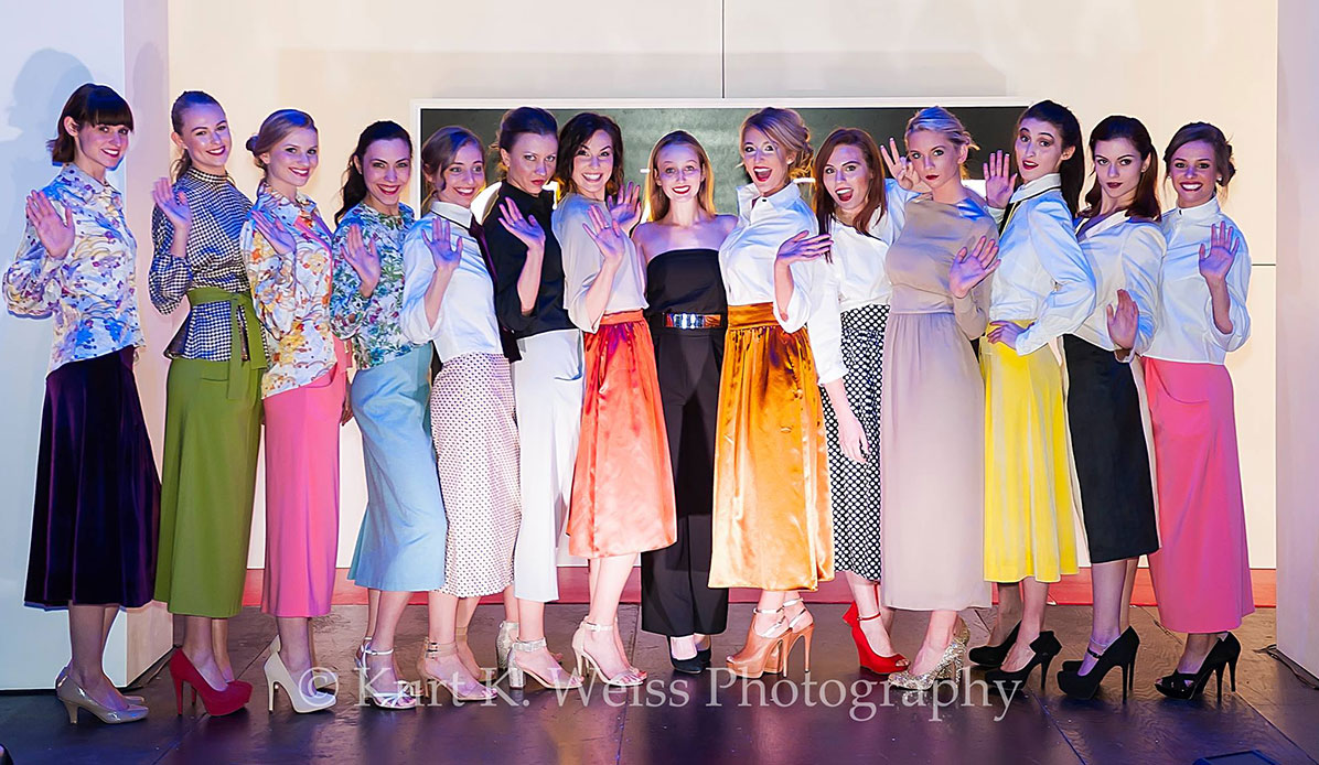 Fourteen models wave to the camera after a fashion show.